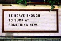 a white brick wall with a written mural in black all-caps type "Be brave enough to suck at something new."